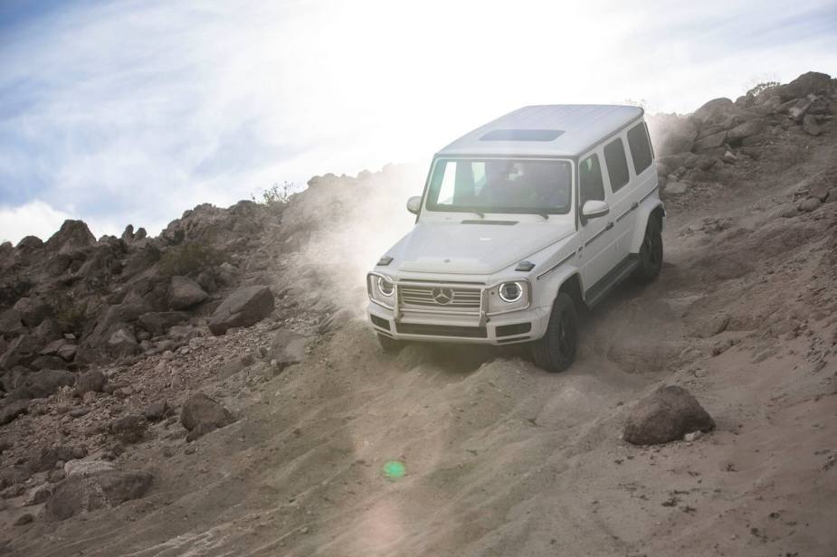 A white Mercedes-Benz G Wagon descends a sandy off-road trail, boulders visible on either side of the 4WD SUV.