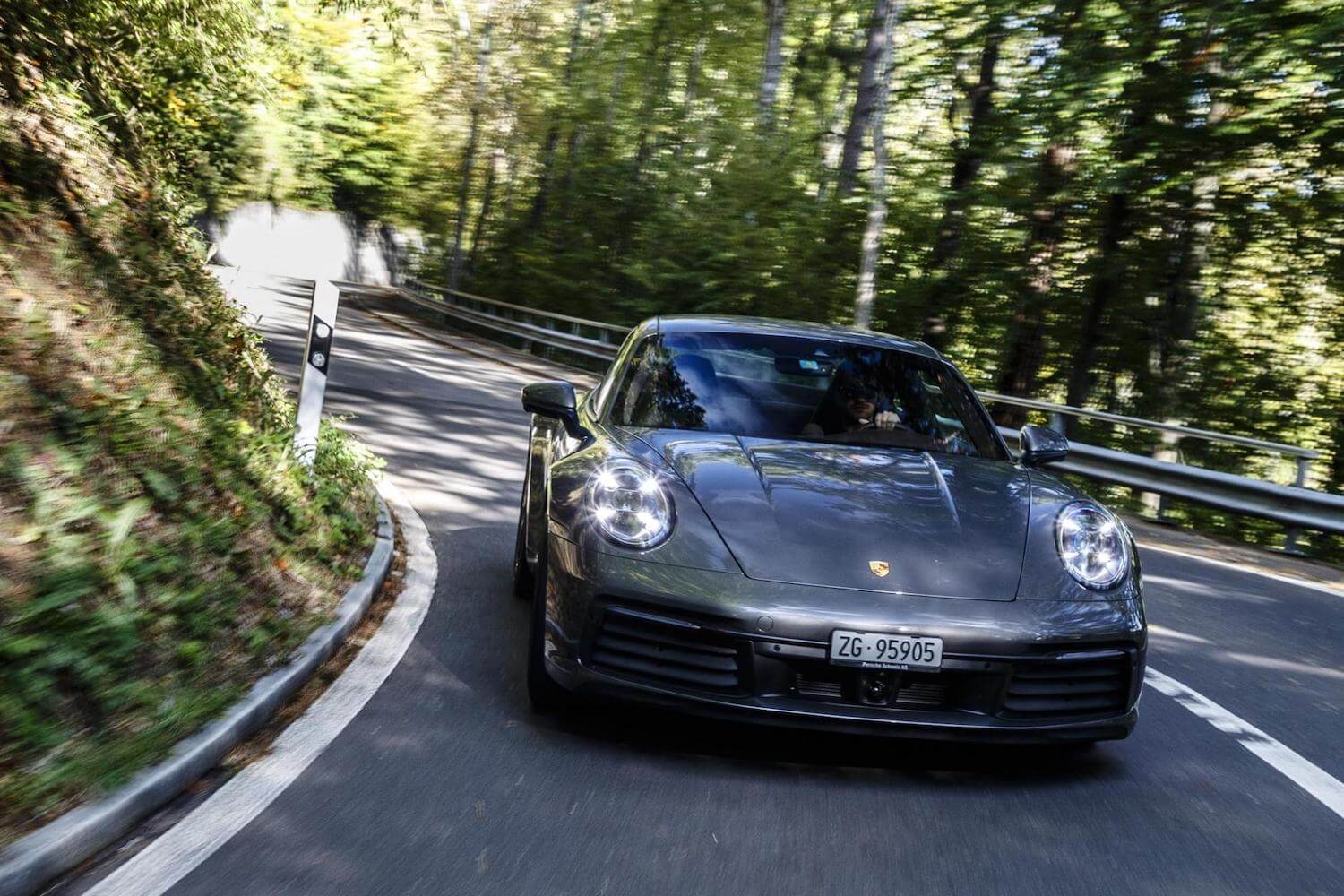 A gray Porsche 911 sports car races around a corner in a mountain road and towards the camera, trees visible in the background.