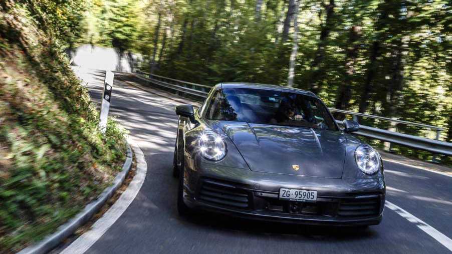 A gray Porsche 911 sports car races around a corner in a mountain road and towards the camera, trees visible in the background.