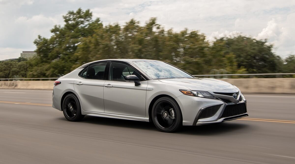 A gray Toyota Camry Hybrid cruises down a back road showing off its side profile.