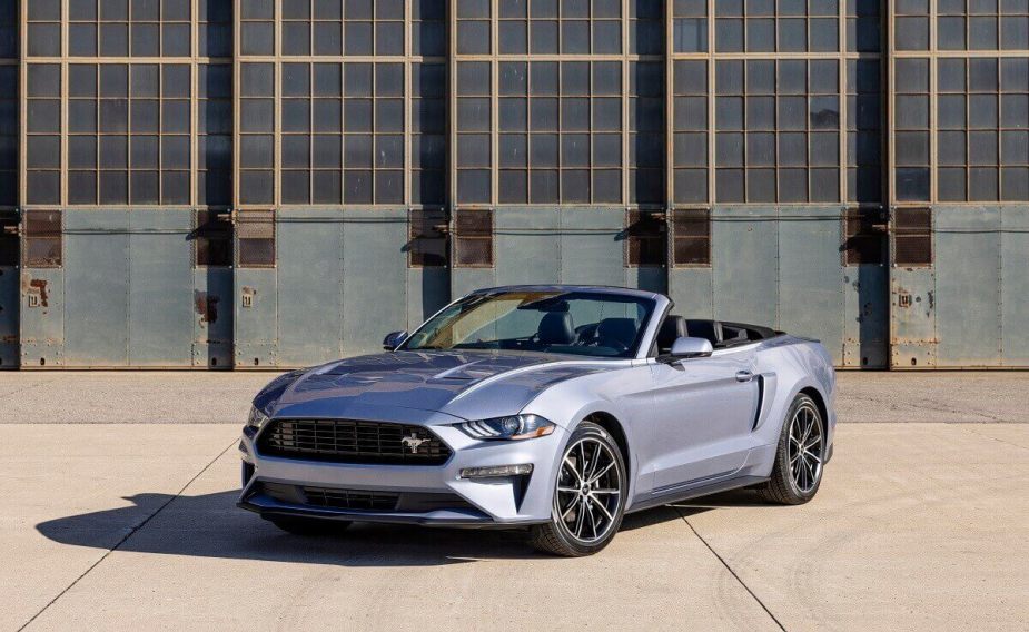 A 2023 Ford Mustang EcoBoost shows off its convertible top and silver paint work in front of a hangar. 