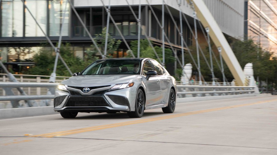 A Toyota Camry Hybrid shows off its silver paintwork while it cruises across a bridge in a city.