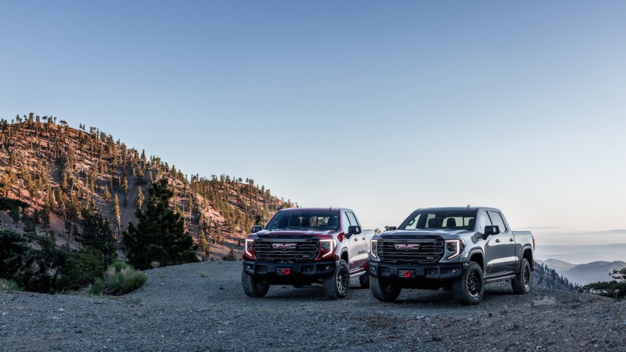 Two 2023 GMC Sierra trucks parked outdoors in front of a mountain and body of water.