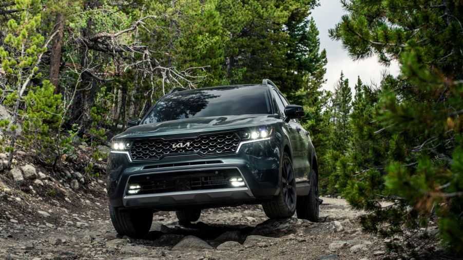 A dark green 2023 Kia Sorento X-Line compact SUV model driving on a gravel dirt road in a forest