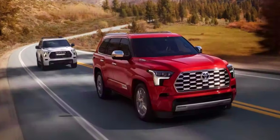 A red 2023 Toyota Sequoia full-size SUV and a white 2023 Toyota Sequoia full-size SUV are driving on the road. 