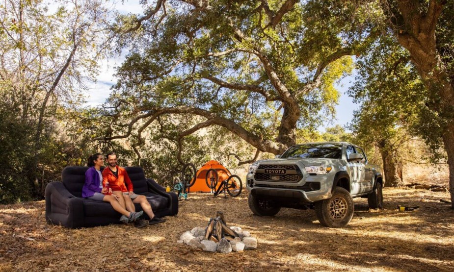 A couple sits by a campfire in the woods, their Toyota Tacoma and tent visible in the background.