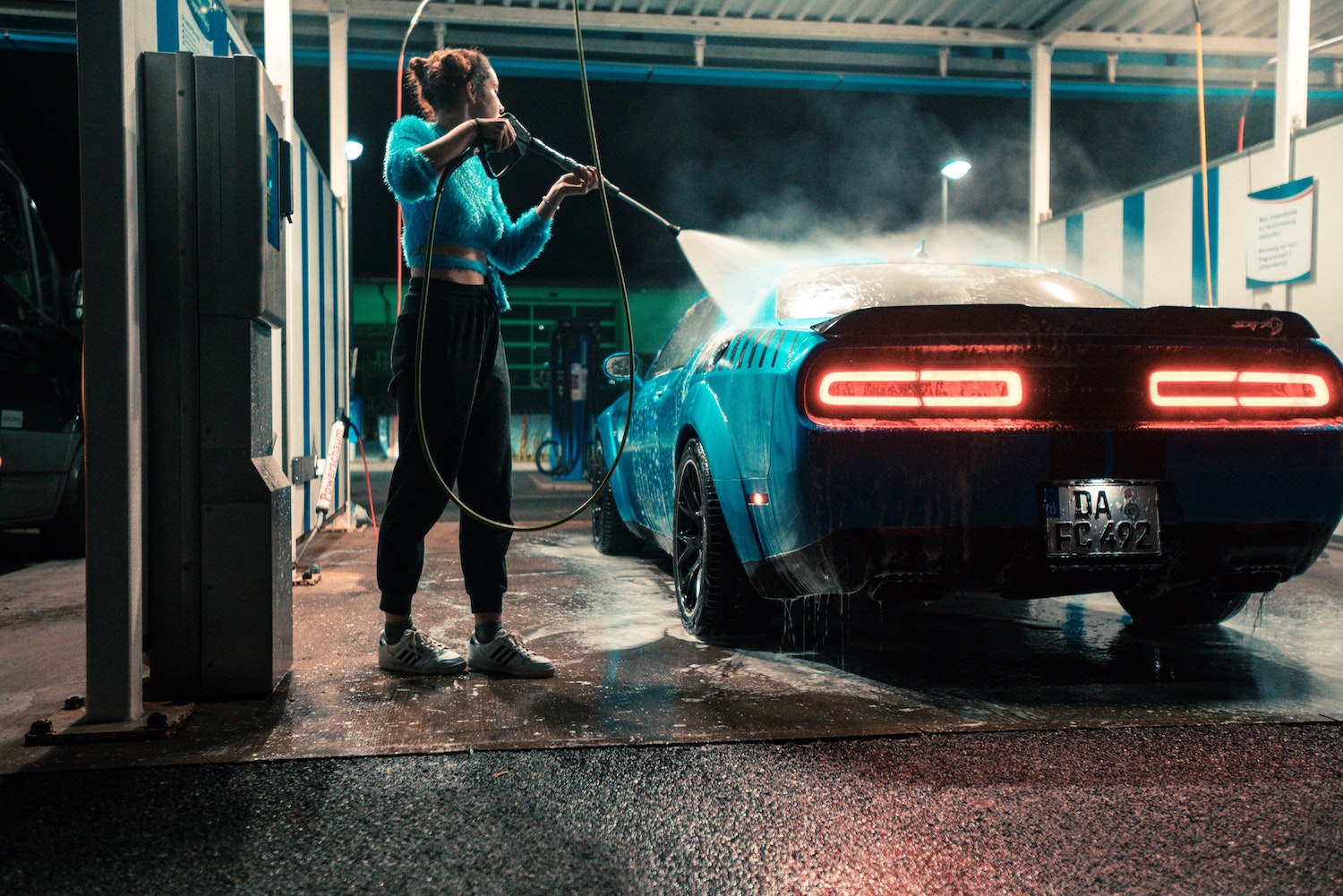 A woman uses a carwash spray gun to clean off a Dodge Challenger sports car at night.