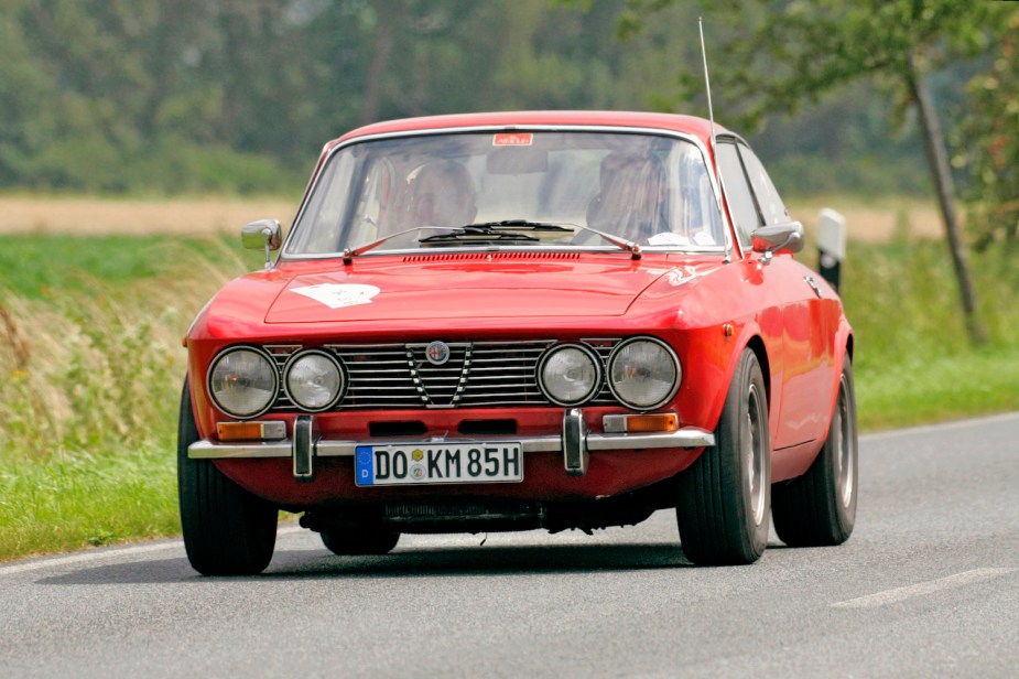 A red 1975 Alfa Romeo coupe driving down a road with grass and trees visible in the background.