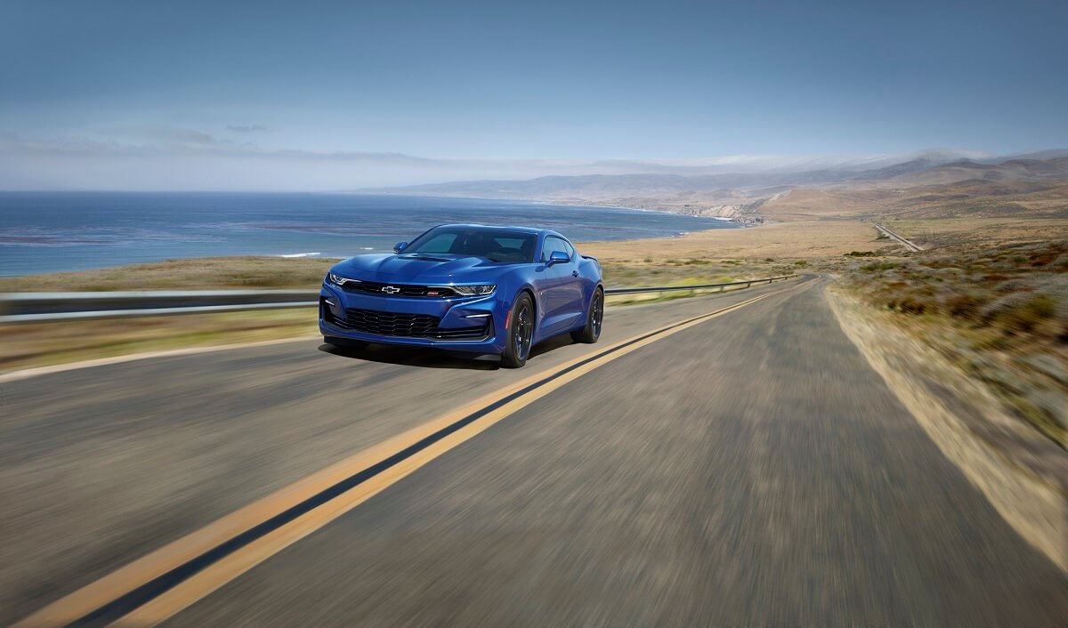 A blue manual transmission Chevrolet Camaro SS, a rival to the Ford Mustang GT, is driving along a lakeside road.