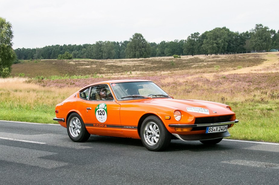 Orange Datsun 240Z coupe parked on a road with trees and fields visible in the background.