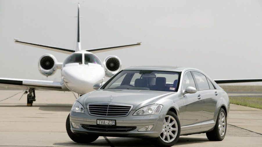 A used silver Mercedes-Benz S-Class luxury car poses in front of a private jet airplane.