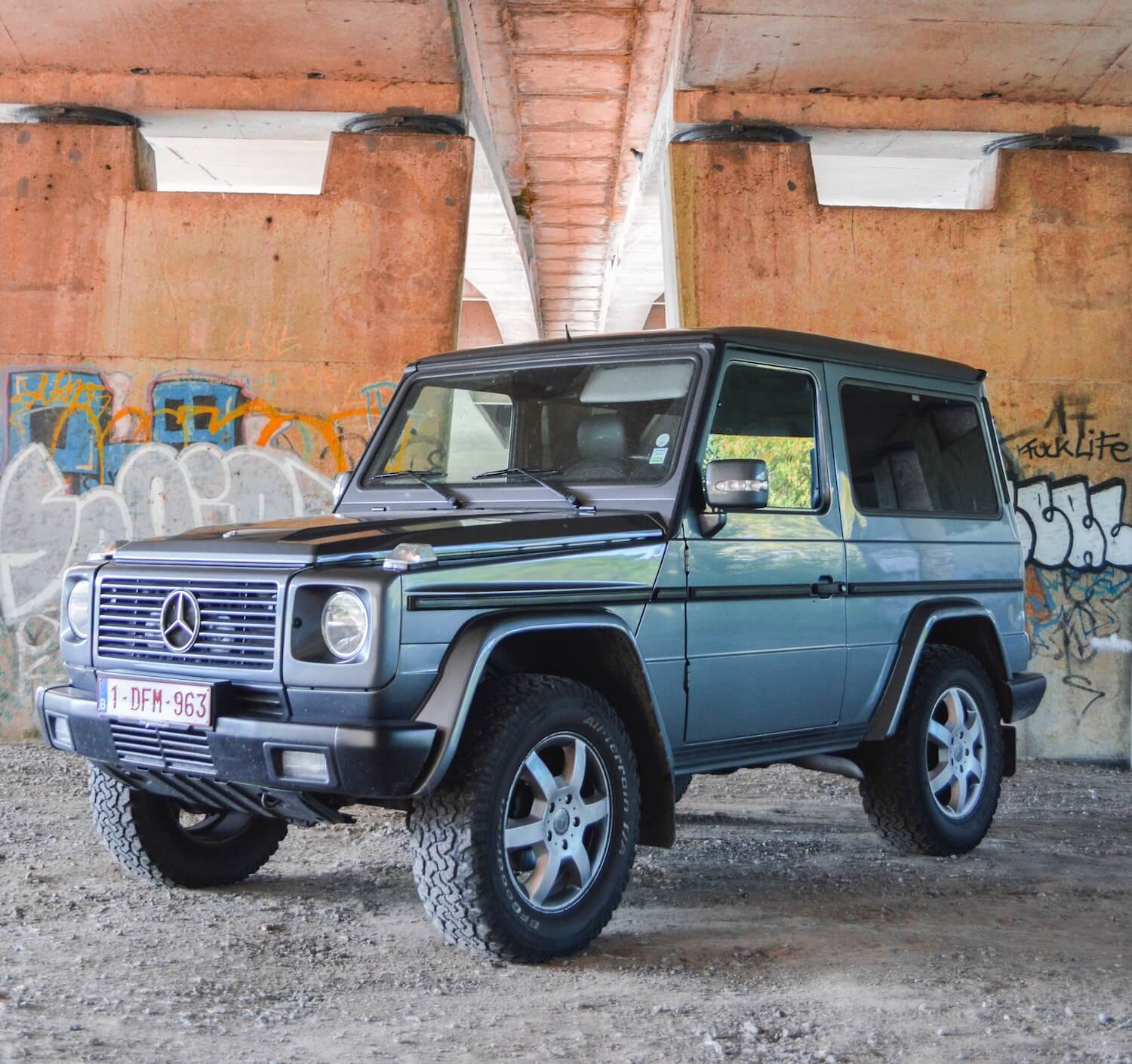 A two-door, used Mercedes-Benz G wagon in blue, parked in front of a wall of graffiti.