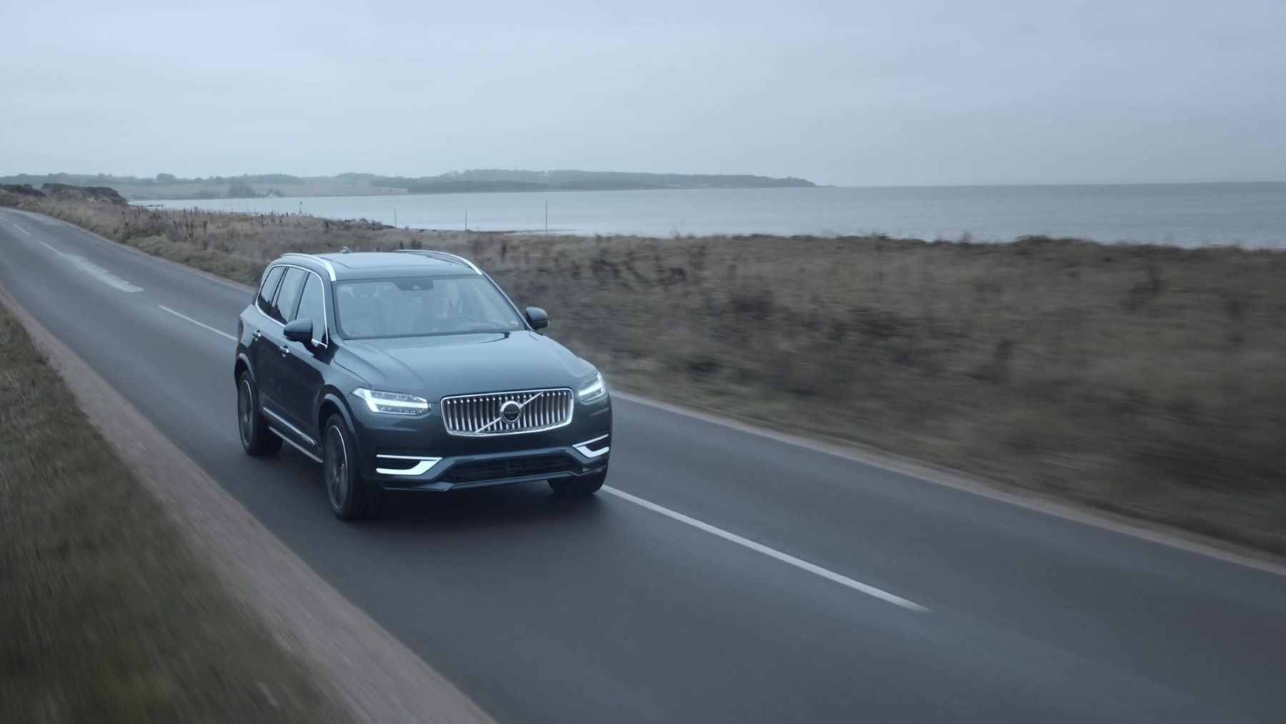 A Volvo XC90 Recharge luxury midsize SUV model in Denim Blue driving past the sea in a fog
