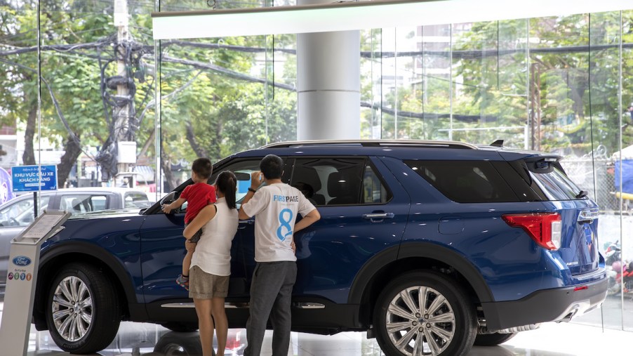 A blue SUV with a family looking at it in a dealership, potentially wonder why SUVs are so expensive.