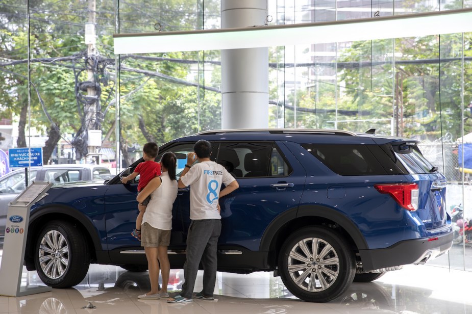 A blue SUV with a family looking at it in a dealership, potentially wonder why SUVs are so expensive.