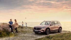 A bronze/copper Mercedes-Benz EQB luxury SUV model parked on a gravel road near a sunset beach as a family plays