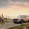 A bronze/copper Mercedes-Benz EQB luxury SUV model parked on a gravel road near a sunset beach as a family plays