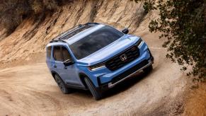 A light blue 2023 Honda Pilot TrailSport midsize crossover SUV model driving off-road on dirt trails