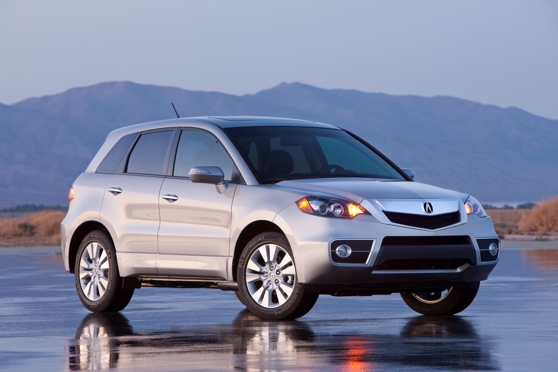 A silver-gray 2010 Acura RDX compact luxury crossover SUV model parked on a wet asphalt lot