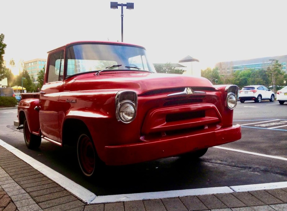 A red International Harvester step-side classic pickup truck parked in a lot, crossovers visible in the background.
