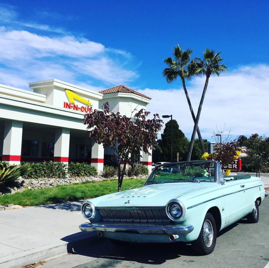 A light blue convertible slant-six Dodge Dart parked in front of an in-and-out burger and a pair of palm trees in Redwood City California.