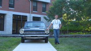 A young man standing next to a classic 1964 Dodge Dart coupe powered by a slant-six, a building and trees bisible in the background.