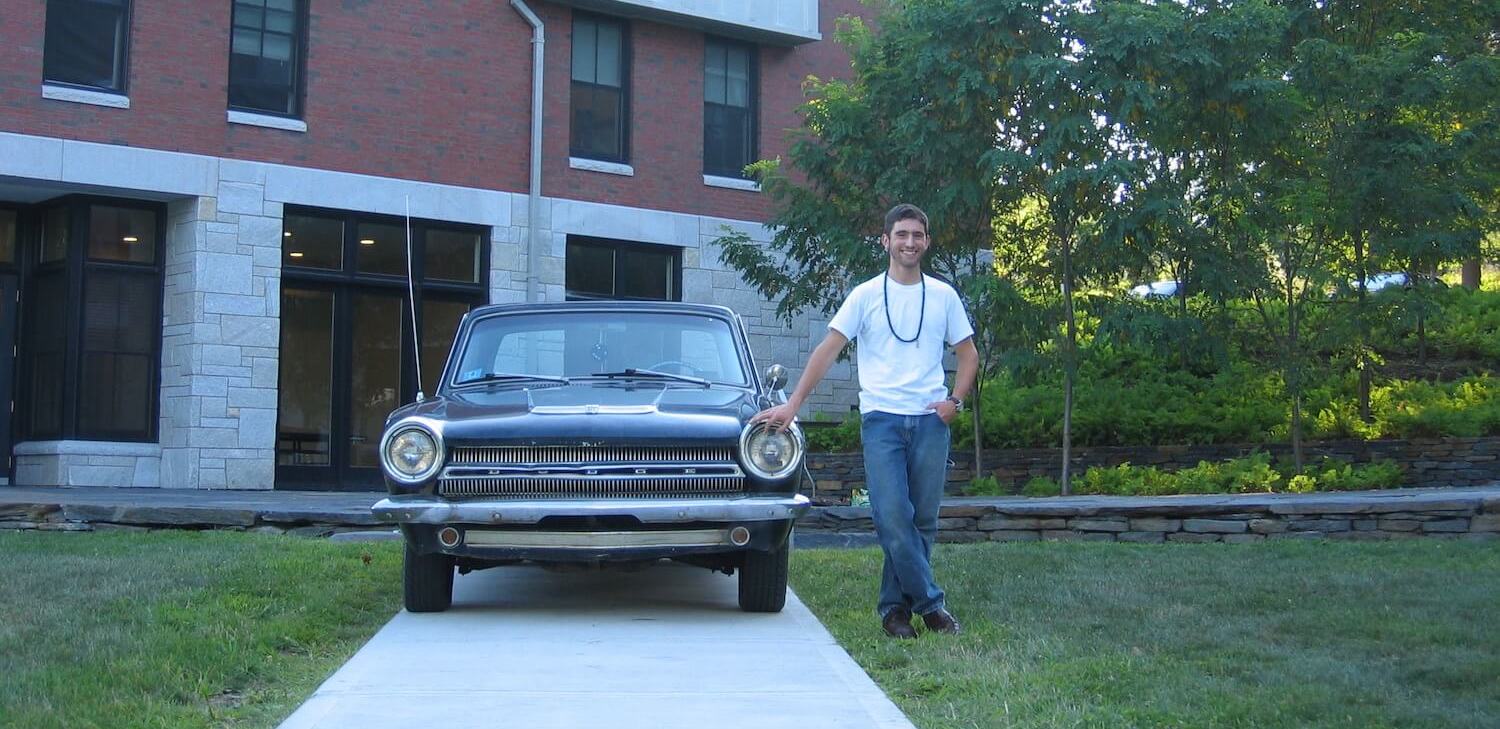 A young man standing next to a classic 1964 Dodge Dart coupe powered by a slant-six, a building and trees bisible in the background.