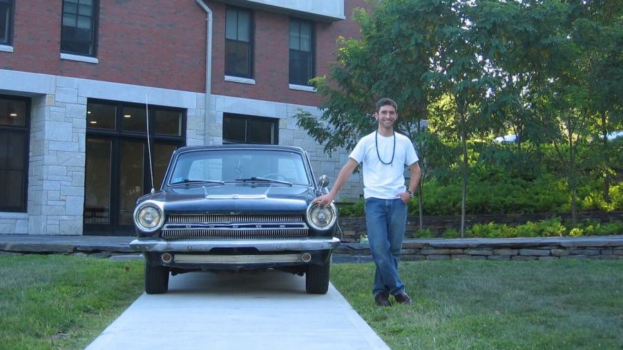 A young man standing next to a classic 1964 Dodge Dart coupe powered by a slant-six, a building and trees bisible in the background.