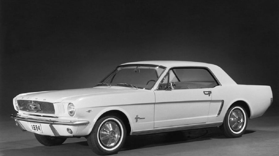 A first-generation 1965 Ford Mustang, what fans call the 1964 1/2 model, shows off its white paintwork and Ford Falcon platform on a stage.