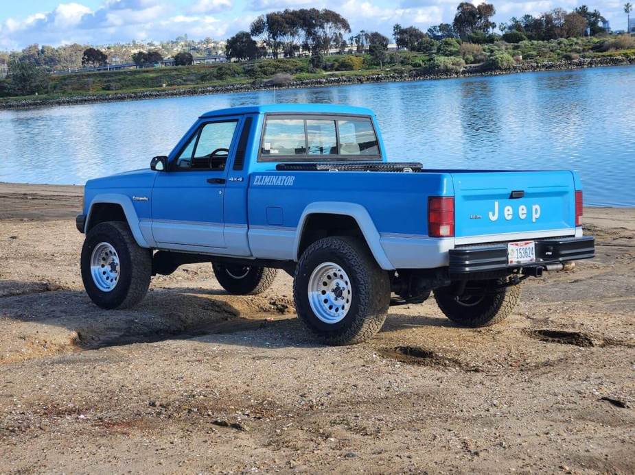 Blue 1990 Jeep Comanche pickup truck parked on rocks by the water.