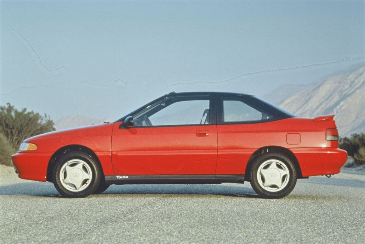 A side profile shot of a red 1994 Hyundai Scoupe model parked near foggy mountains
