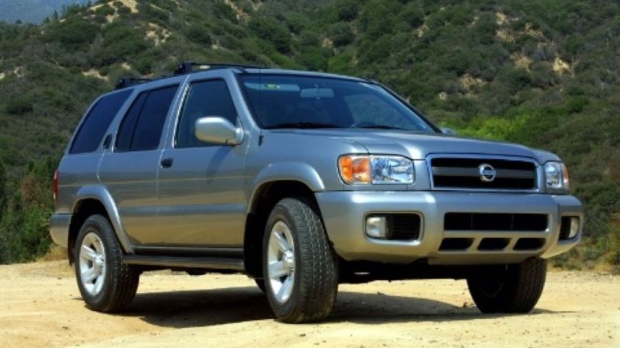 A silver-gray 2003 Nissan Pathfinder midsize SUV model parked on a dirt plain in a mountain forest