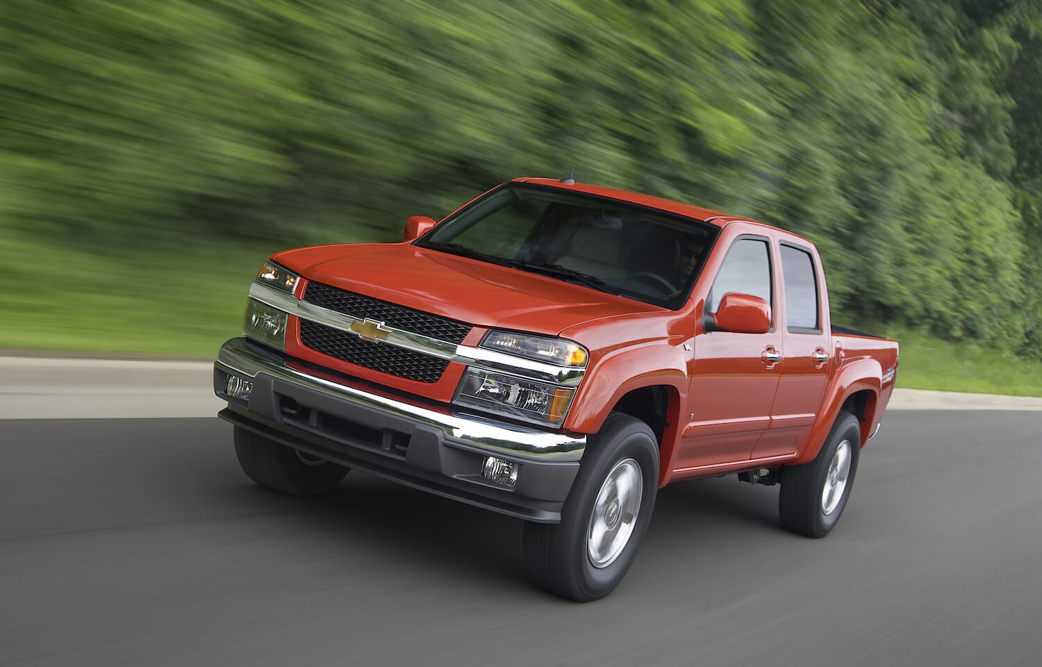An orange 2009 Chevrolet Colorado (first-generation) driving towards the camera fro a promo photo, trees visible in the background.