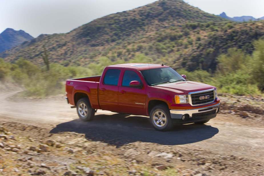A red GMC Sierra pickup truck driving up a dirt road, trees and a mountain ridgeline visible in the background.