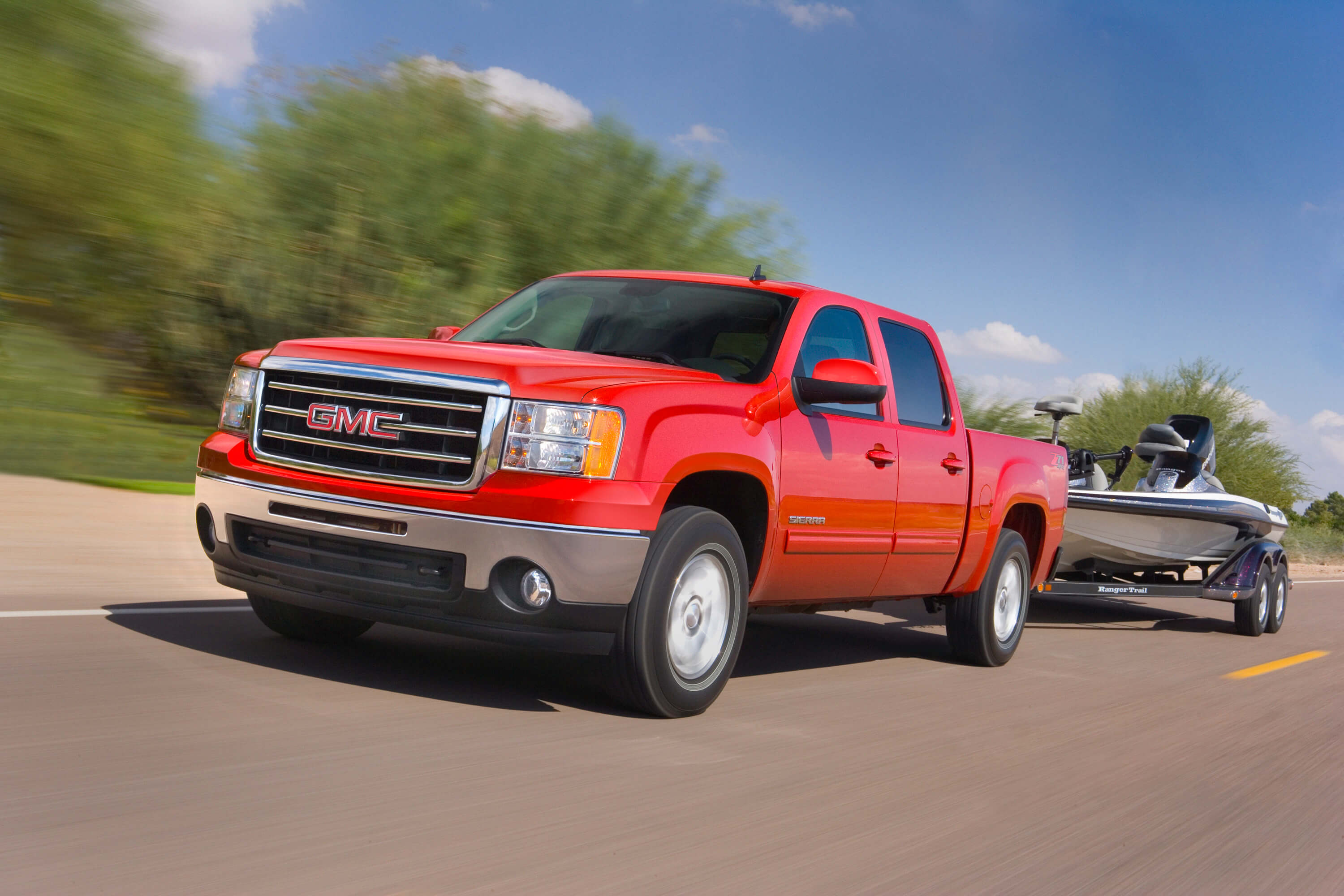 Promo photo of a red 2013 GMC Sierra 1500, a truck with relatively few engine issues, towing a boat down a rural road with trees in the background.
