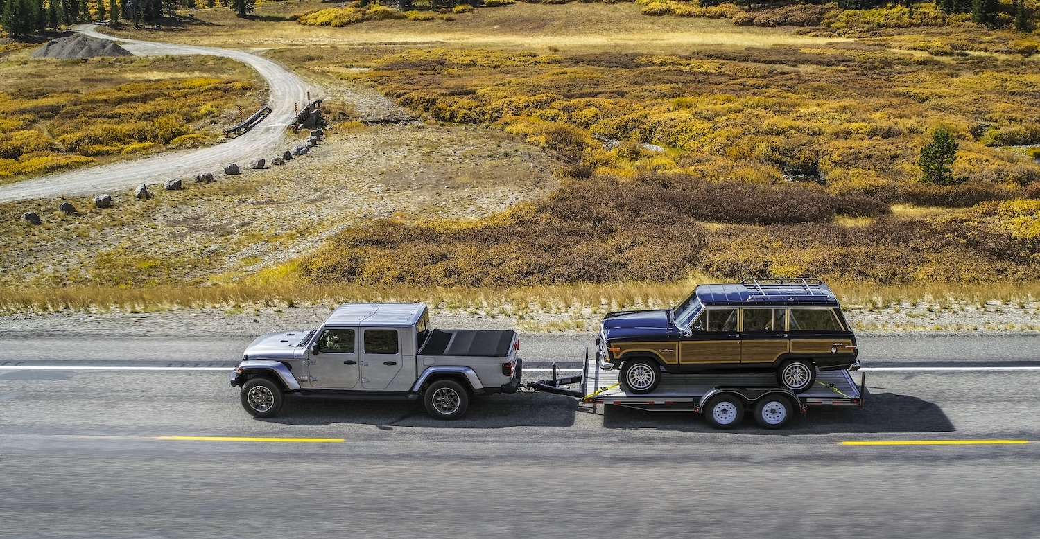 Silver Jeep Gladiator towing a classic Wagoneer SUV, a field visible in the background.