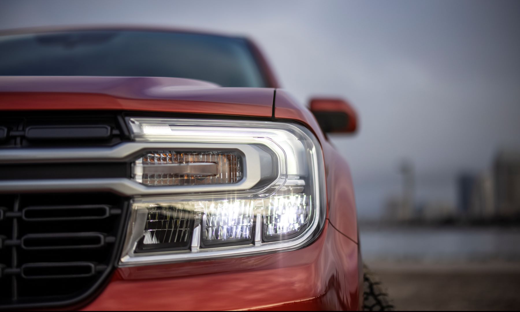 A closeup shot of a red-orange 2022 Ford Maverick Lariat compact pickup truck's headlights and grille
