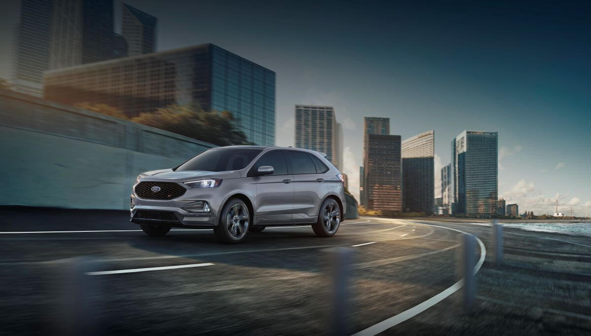 A dark gray 2023 Ford Edge midsize SUV model driving on a highway within an urban city