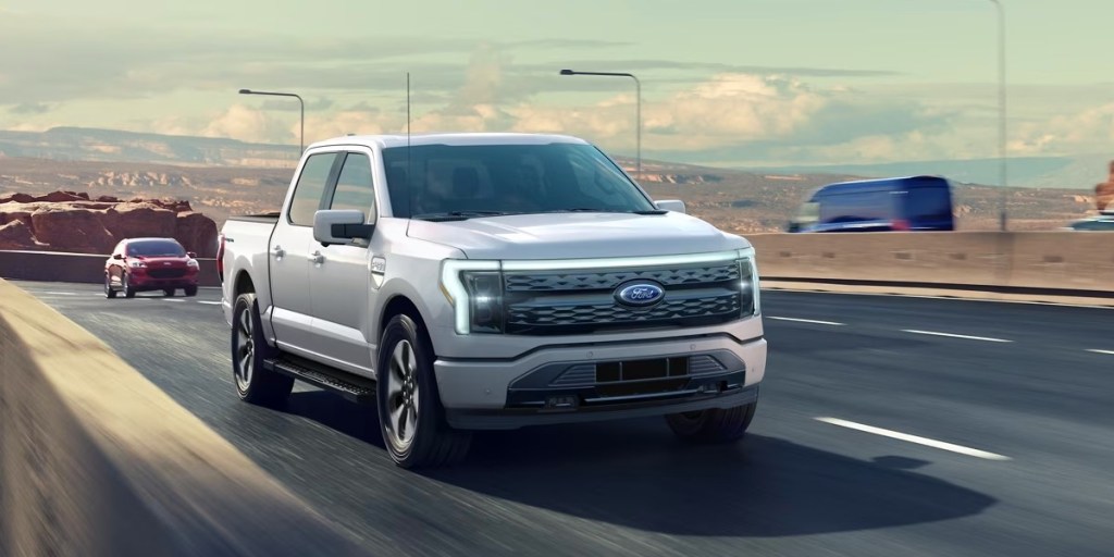 A white 2023 Ford F-150 Lightning electric pickup truck is driving on the road.