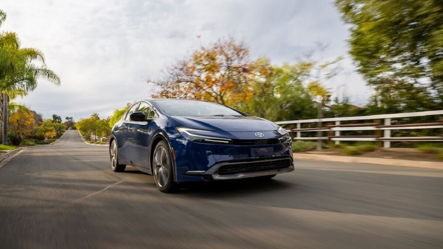 A blue Toyota Prius cruises the countryside on a one-car road.