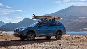 A blue 2023 Subaru Outback, which is a midsize SUV best for you, driving outdoors in front of a mountain and body of water.