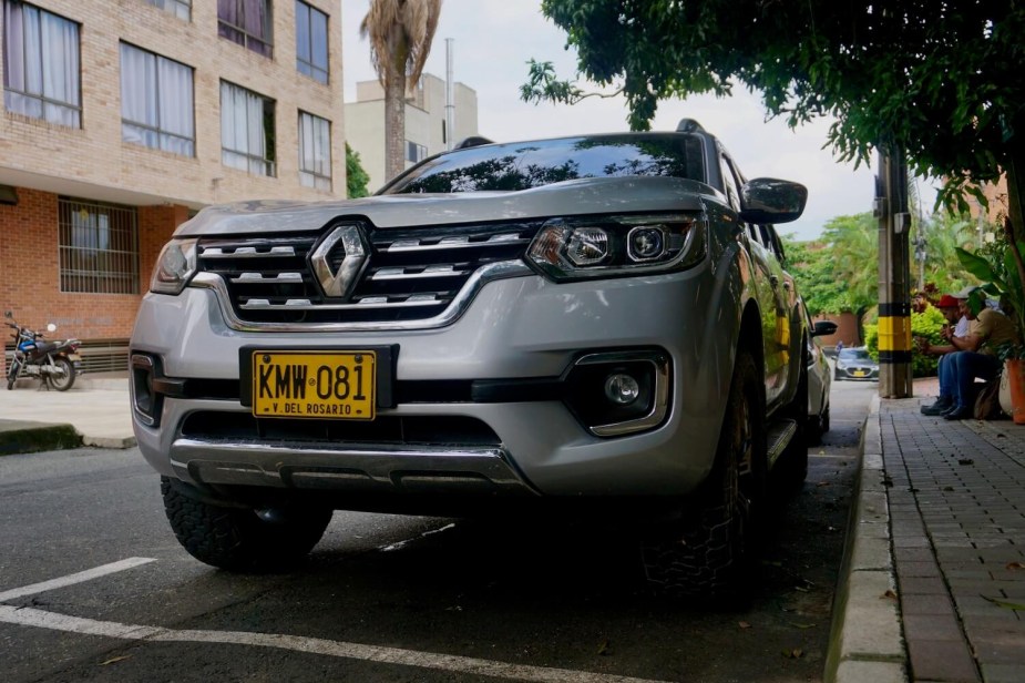 The grille of a silver Renault Alaskan midsize pickup truck wearing Colombian plates, trees and apartment buildings visible in the background.