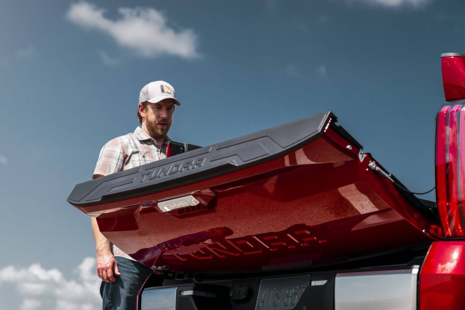 A man raises the tailgate on a red 2023 Toyota Tundra full-size pickup truck, blue sky visible in the background.