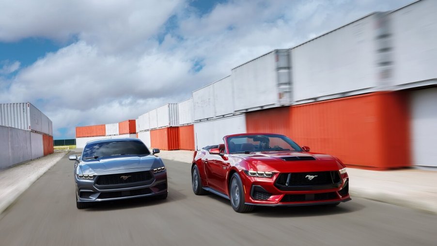 A set of new 2024 Ford Mustangs, including a GT, blast through a harbor showing off their gray-blue and red paint schemes.