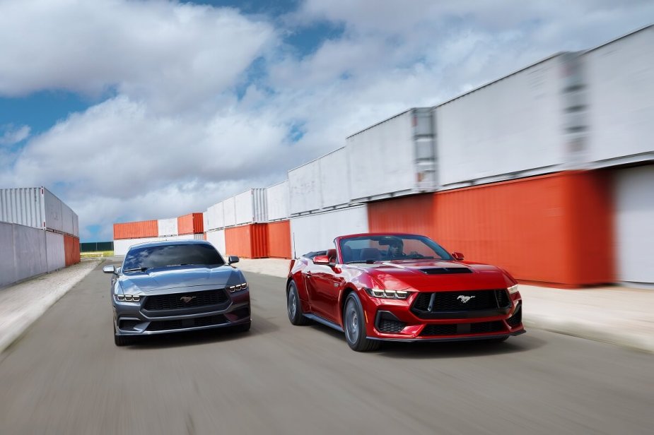 A set of new 2024 Ford Mustangs, including a GT, blast through a harbor showing off their gray-blue and red paint schemes. 