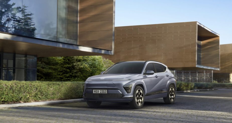 Front three quarter view of a 2024 Hyundai Kona sitting still in front of a building with greenery and late-day shadows. 