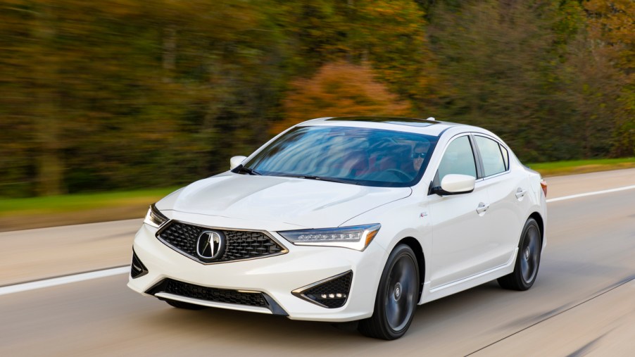 A white Acura ILX driving down the road in a wooded area.
