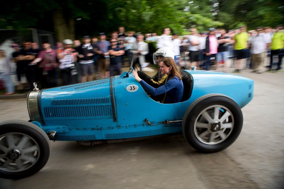 Bugatti Type 35 Grand Prix race car in French racing blue driving towards the course at Goodwood, a blurry crowd visible in the background.