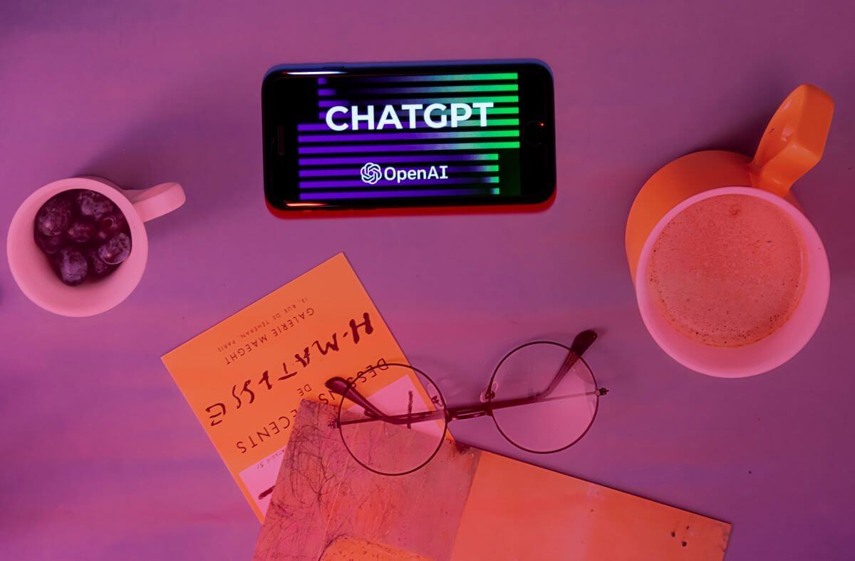 ChatGPT from OpenAI on a smartphone next to coffee, books, and glasses, on a table