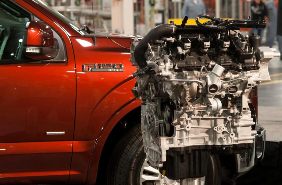An F-150 engine in front of a the fender of a red truck sitting on a factory floor.